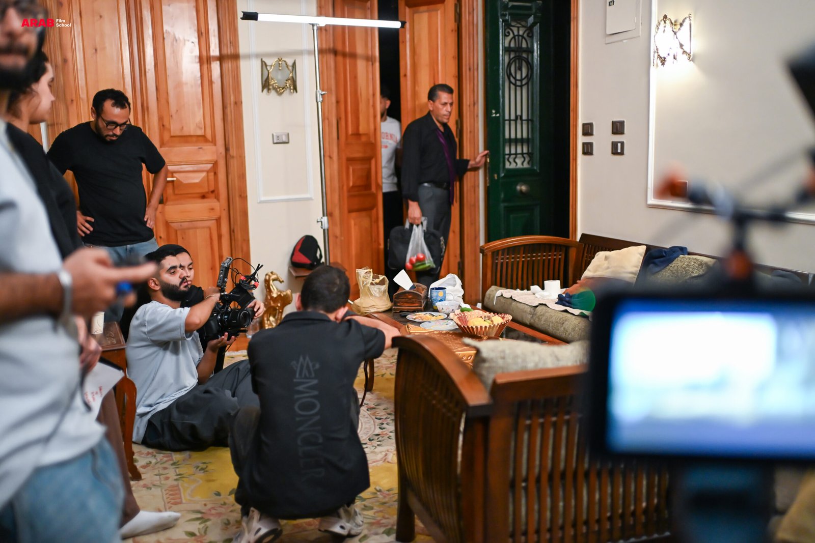 طلاب يصورون مشهداً داخل شقة، والمصور يجلس على الأرض بالكاميرا بينما يدخل الممثل من الباب